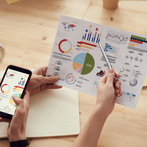 People's Hands Near Printed Analytics Reports, Placed on Light Wood Desk