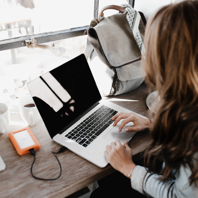 Woman in Denim Jacket Sitting at Coffee Shop Table with MacBook Pro and Cup of Coffee