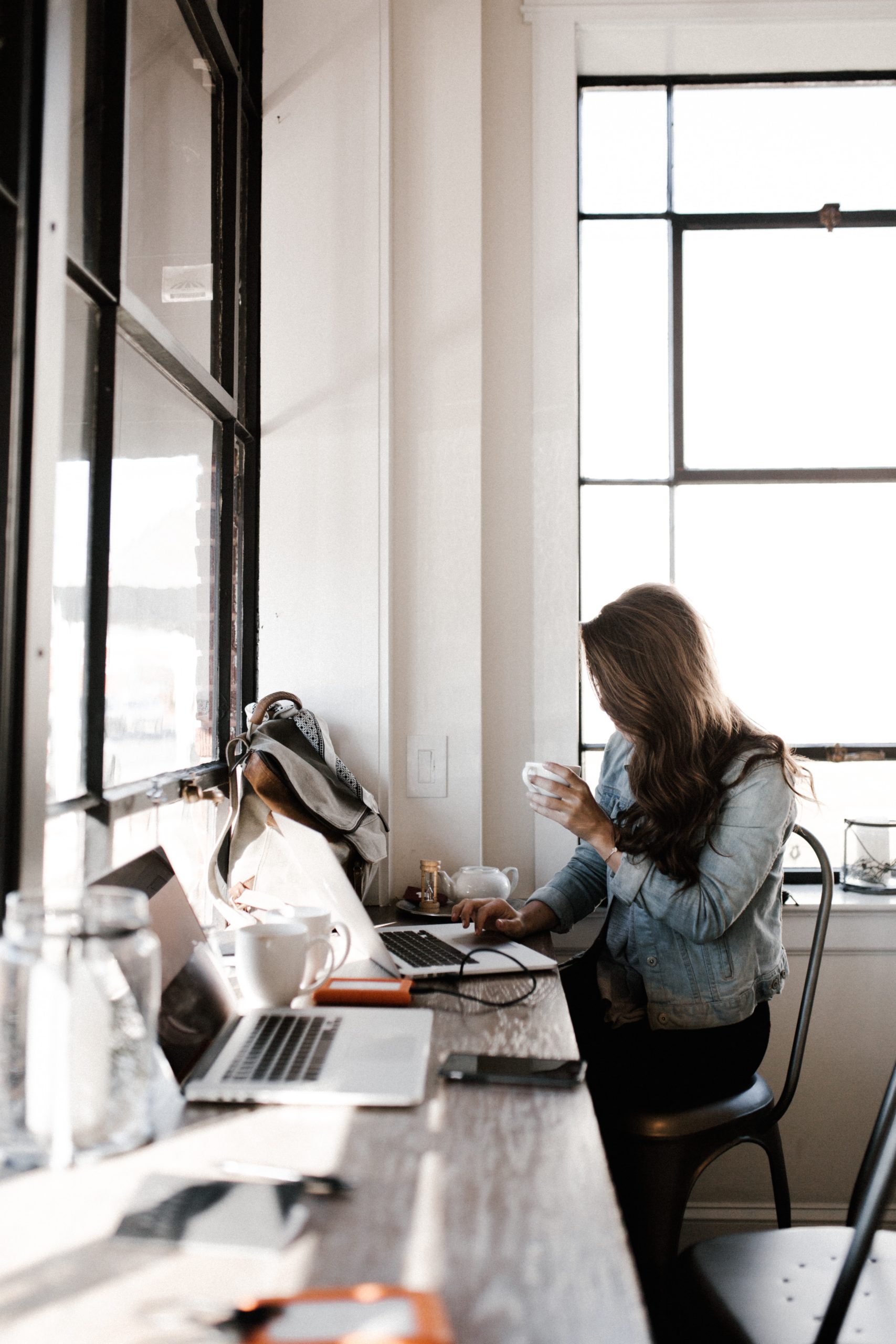 Woman in Denim Jacket Sitting at Coffee Shop Table with MacBook Pro and Cup of Coffee