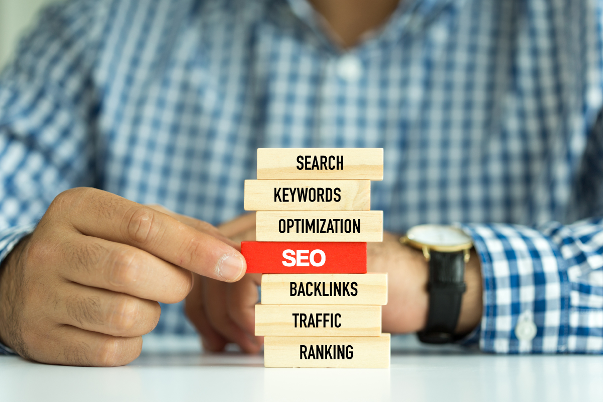 Jenga Blocks with SEO Terms Stacked, Man Wearing Blue Checkered Shirt Sitting Behind